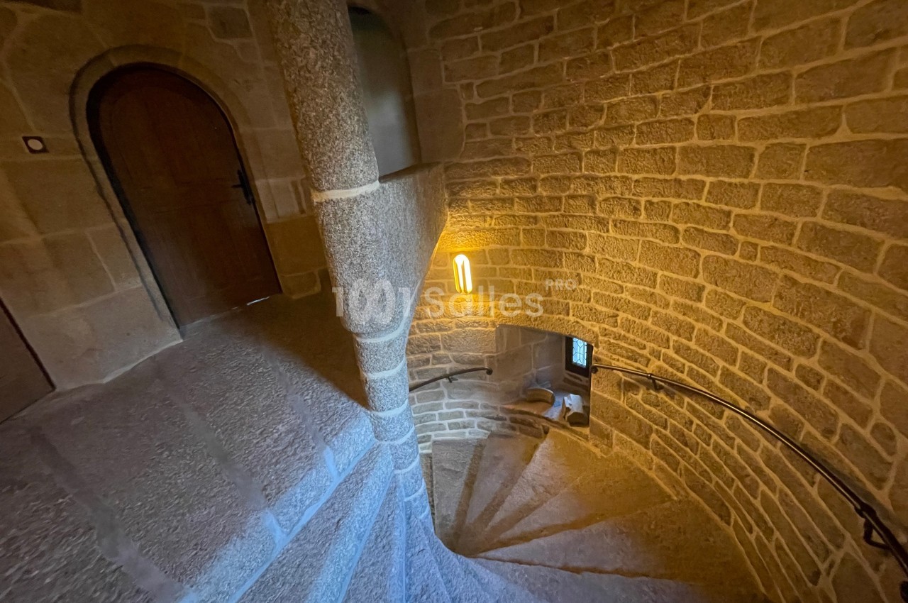 Escalier en colimaçon en pierre éclairé par une lampe murale, avec des murs en pierre apparente.