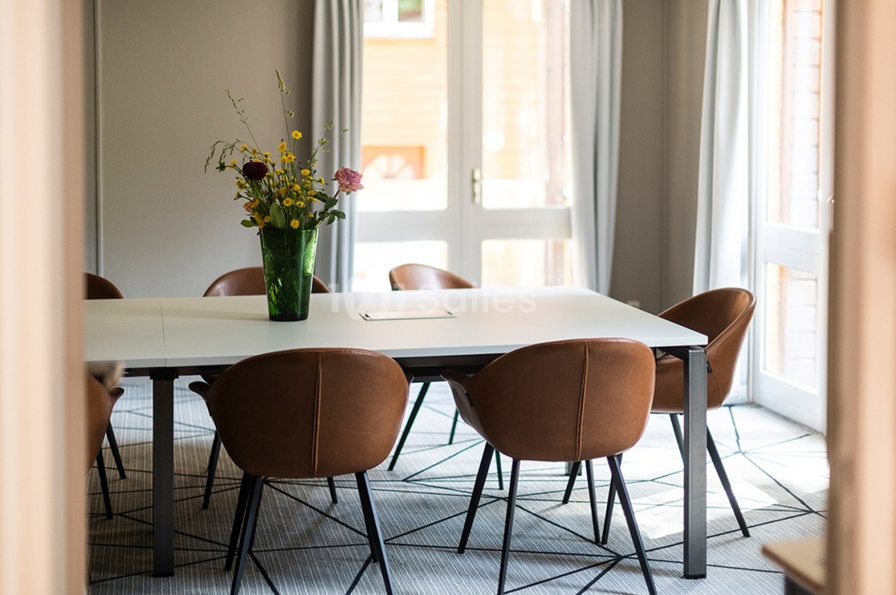 Salle lumineuse avec table blanche, chaises en cuir marron et vase de fleurs colorées au centre.