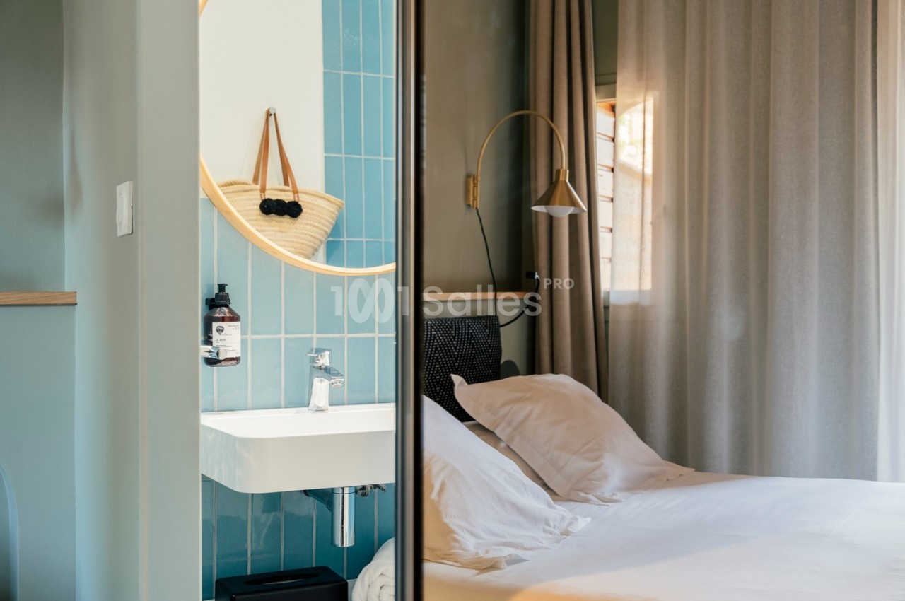 Vue d'une chambre lumineuse avec un lit blanc, un miroir rond au-dessus d'un lavabo et des carreaux bleus.