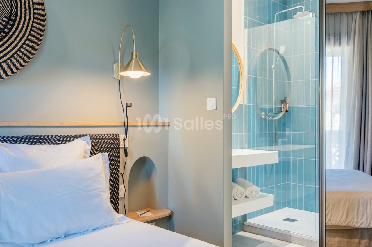 Chambre lumineuse avec lit double, mur bleu, lampe murale et salle de bain attenante aux carreaux bleus.