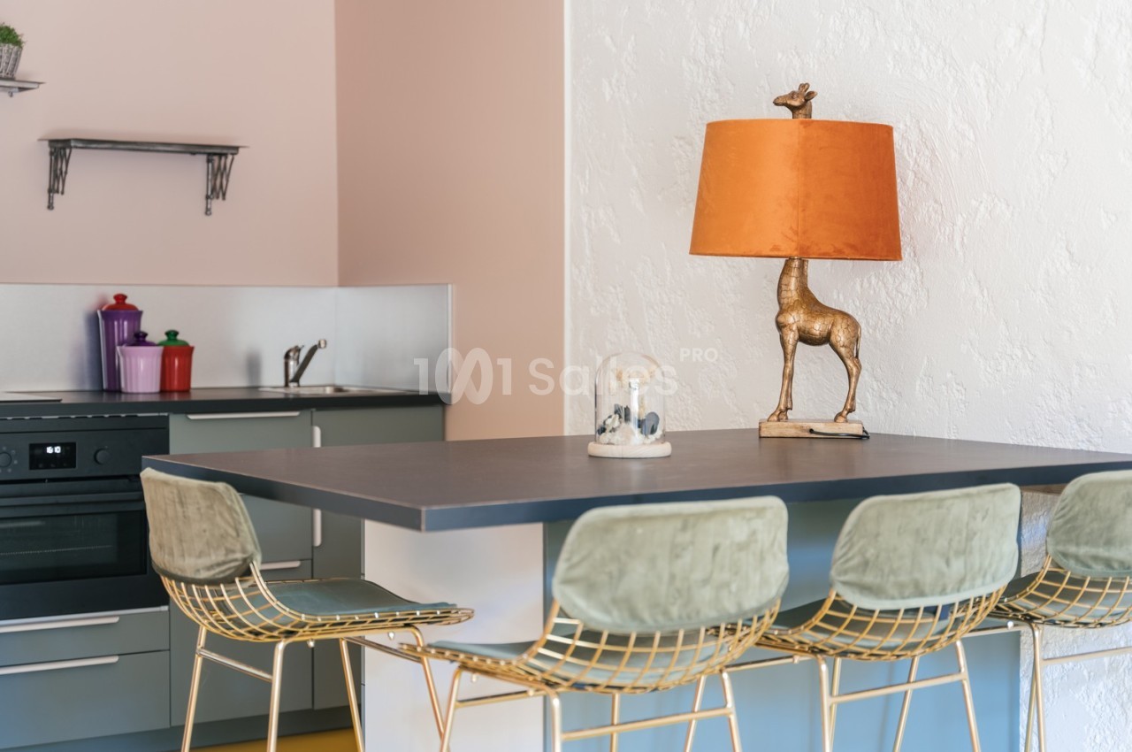 Cuisine moderne avec un plan de travail, des tabourets dorés, une lampe décorative en forme de girafe et des étagères…