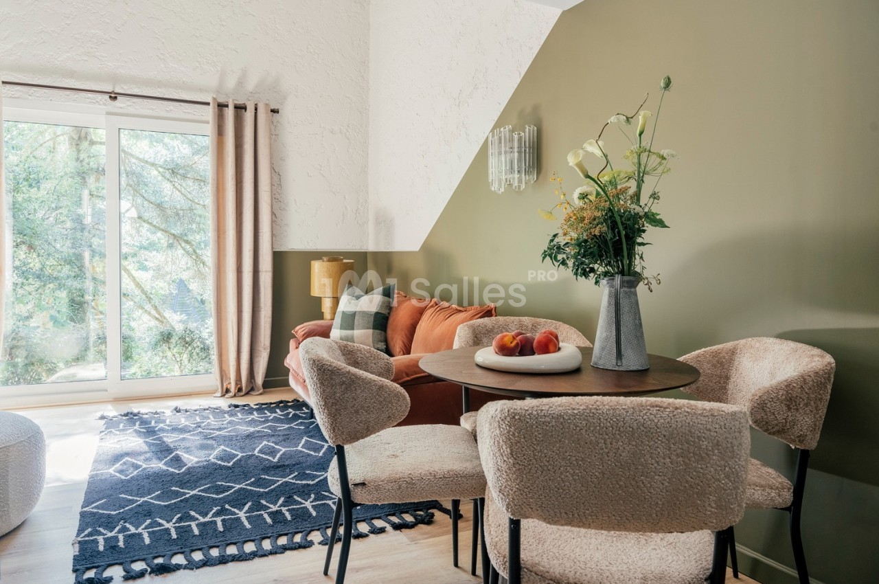 Salle de séjour lumineuse avec table ronde, chaises en tissu, canapé orange, tapis bleu et grande baie vitrée.