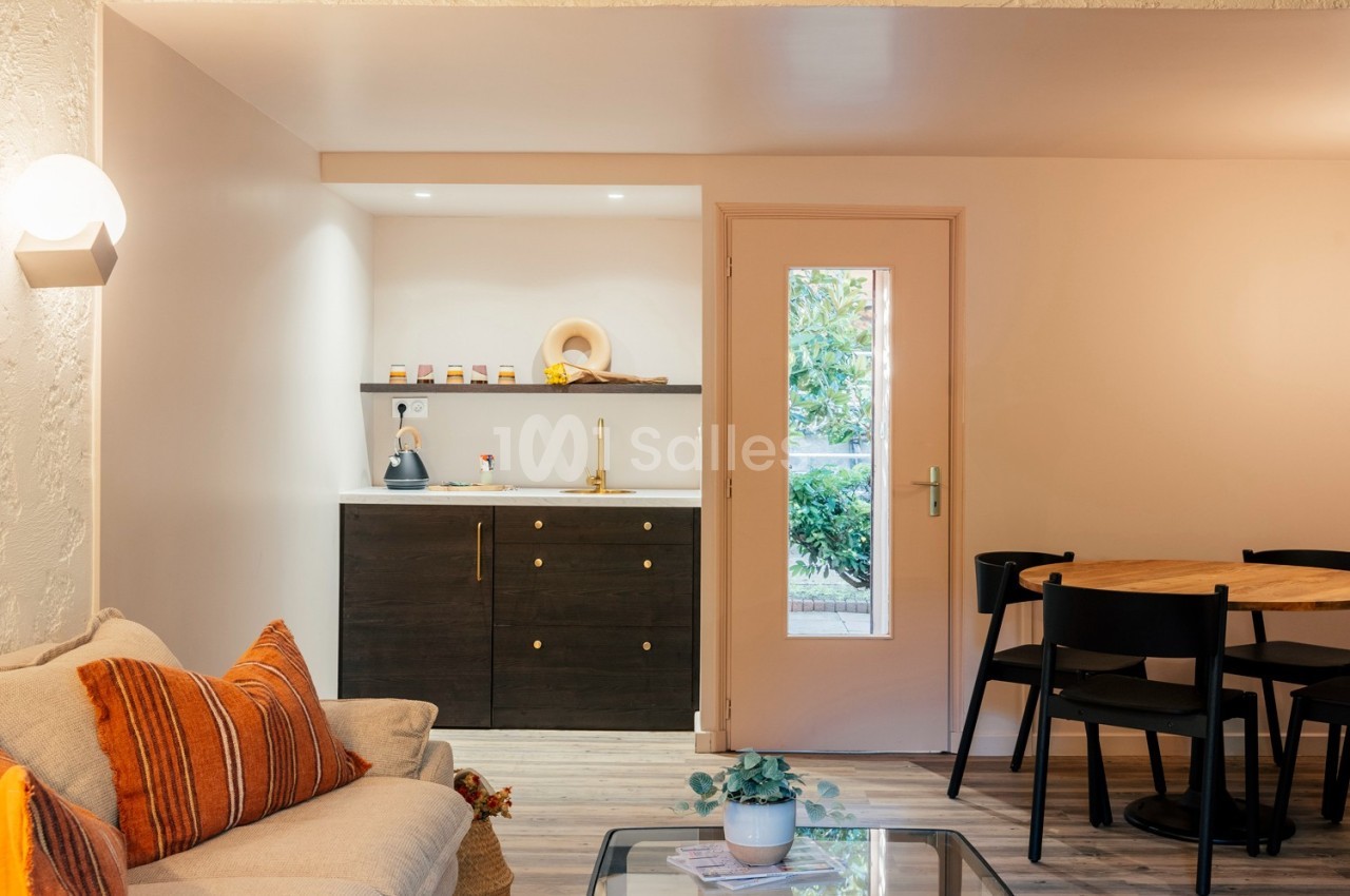 Coin salon avec canapé, table basse et cuisine ouverte équipée, éclairé par une porte vitrée donnant sur l'extérieur.