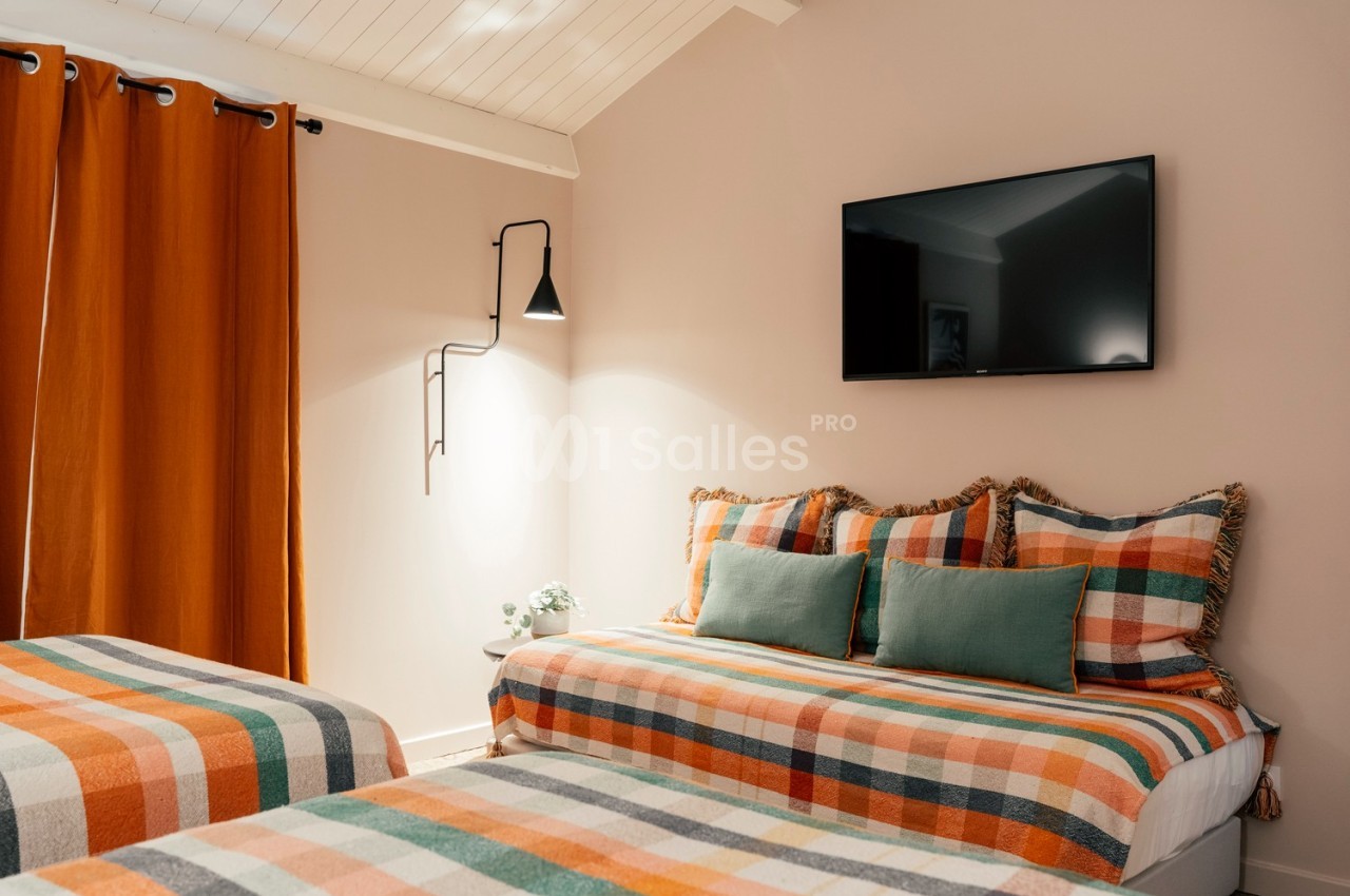 Chambre avec lits et coussins à motifs colorés, rideaux orange, mur beige, téléviseur mural et éclairage moderne.