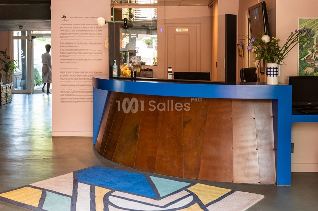 Comptoir d'accueil incurvé en bois et bleu, décoré de fleurs, avec un tapis coloré au sol dans un espace lumineux.