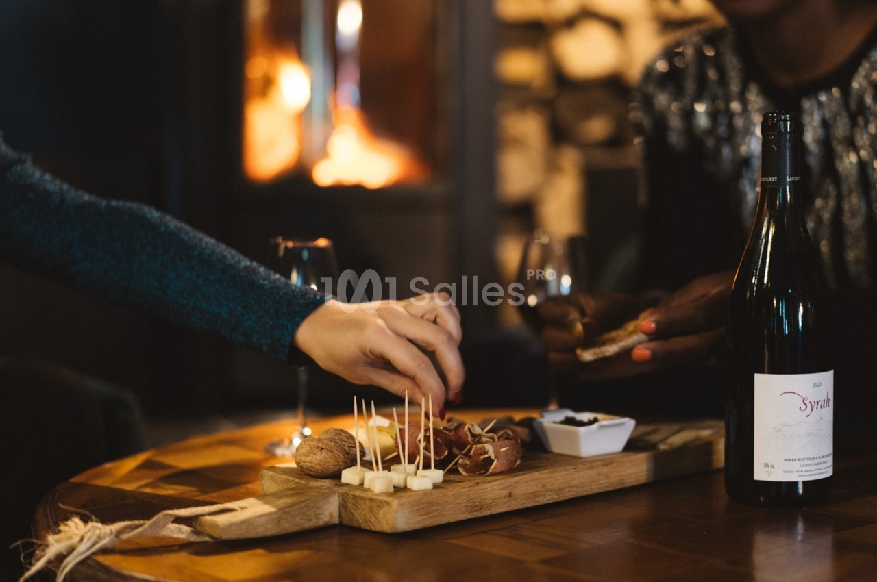 Plateau de fromages et noix sur une table en bois, avec des mains servant et un feu de cheminée en arrière-plan.
