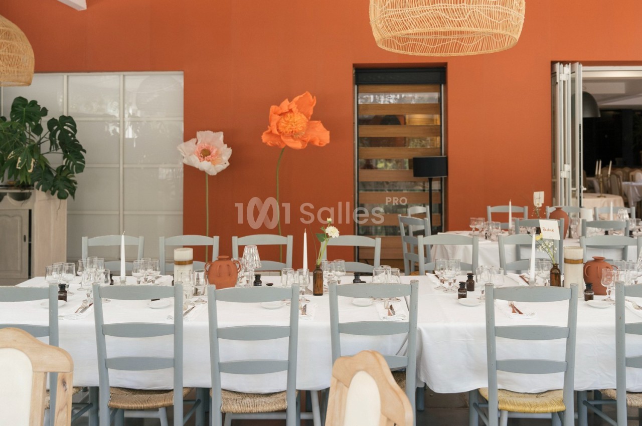 Salle de réception avec tables dressées, nappes blanches, chaises grises et décorations florales devant un mur orange.
