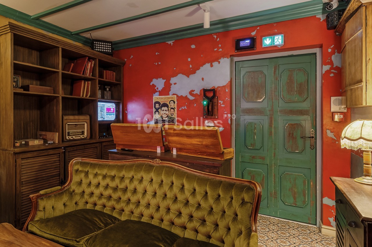 Salon vintage avec canapé vert, meubles en bois, mur rouge usé, porte verte et objets rétro comme une radio et un téléphone.