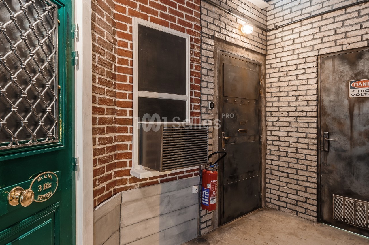 Entrée d'un bâtiment avec murs en briques, porte verte, porte métallique et extincteur fixé au mur.