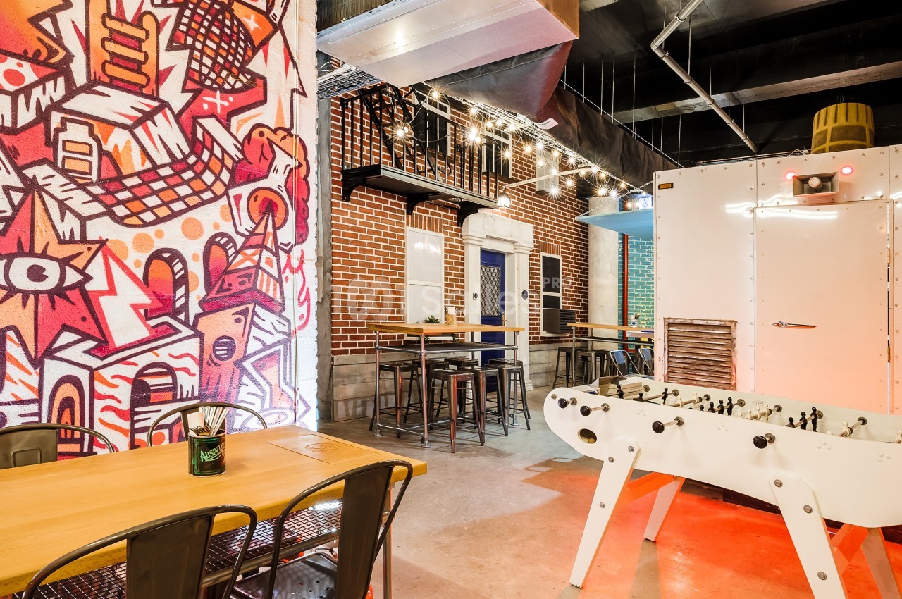 Salle avec décoration urbaine, tables en bois, baby-foot blanc et mur coloré avec graffitis.