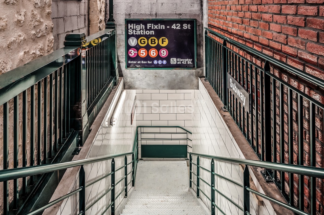 Escalier descendant vers une station de métro, entouré de murs en briques et en pierre, avec un panneau d'indications.