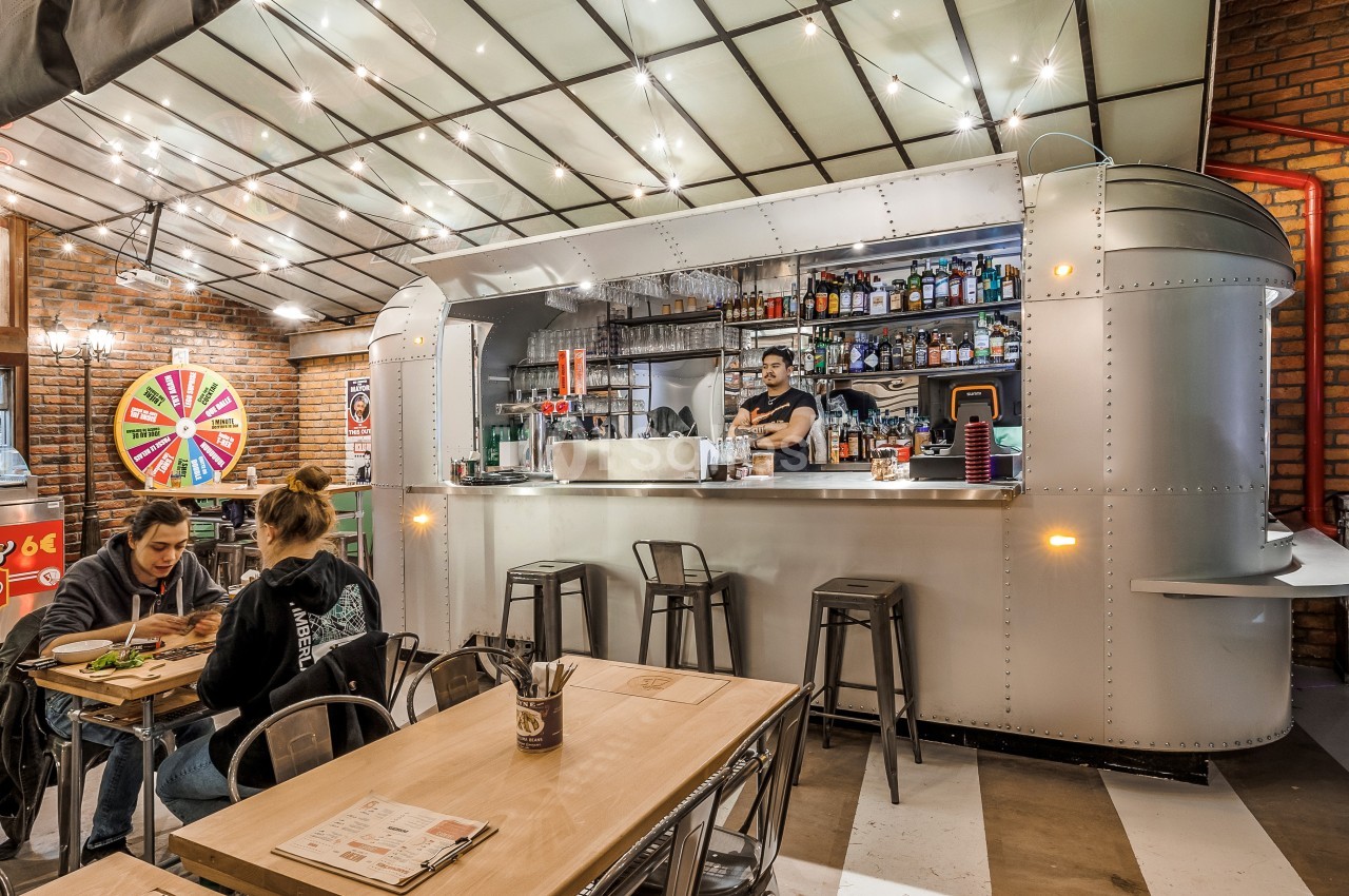 Intérieur d'un restaurant avec un bar en métal, des tables en bois, et un serveur derrière le comptoir.