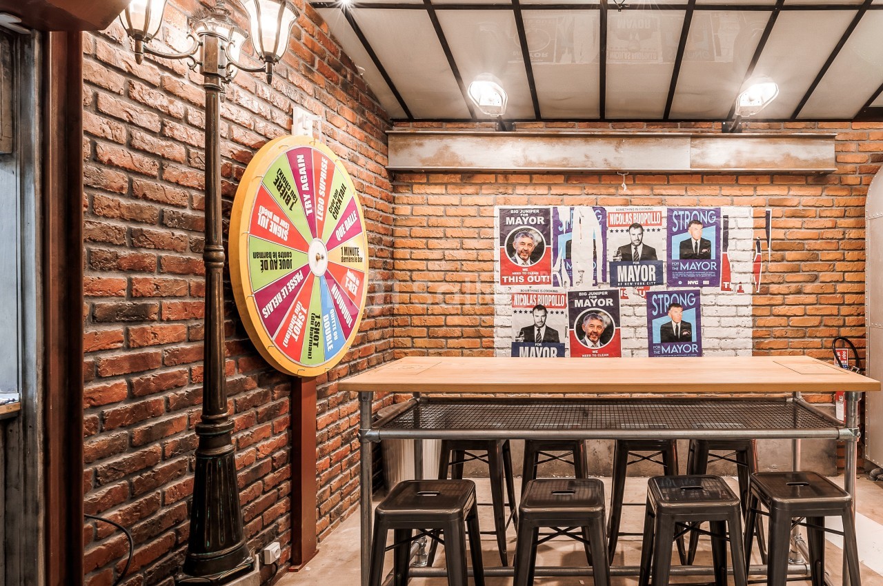 Salle de style industriel avec murs en briques, table haute, tabourets métalliques et roue colorée accrochée au mur.
