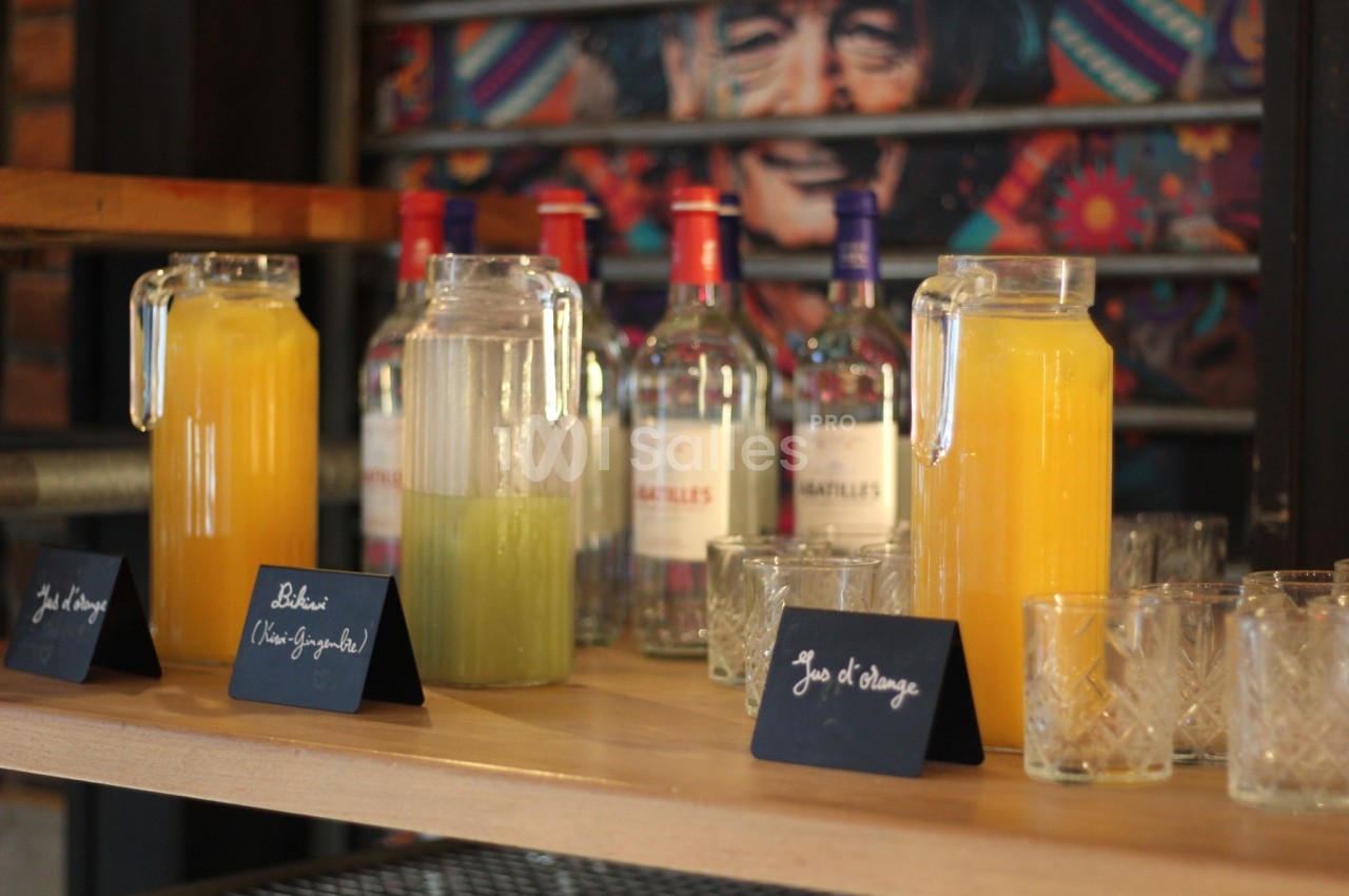 Carafes de jus d'orange et de jus vert alignées sur une étagère, avec des bouteilles d'alcool en arrière-plan.