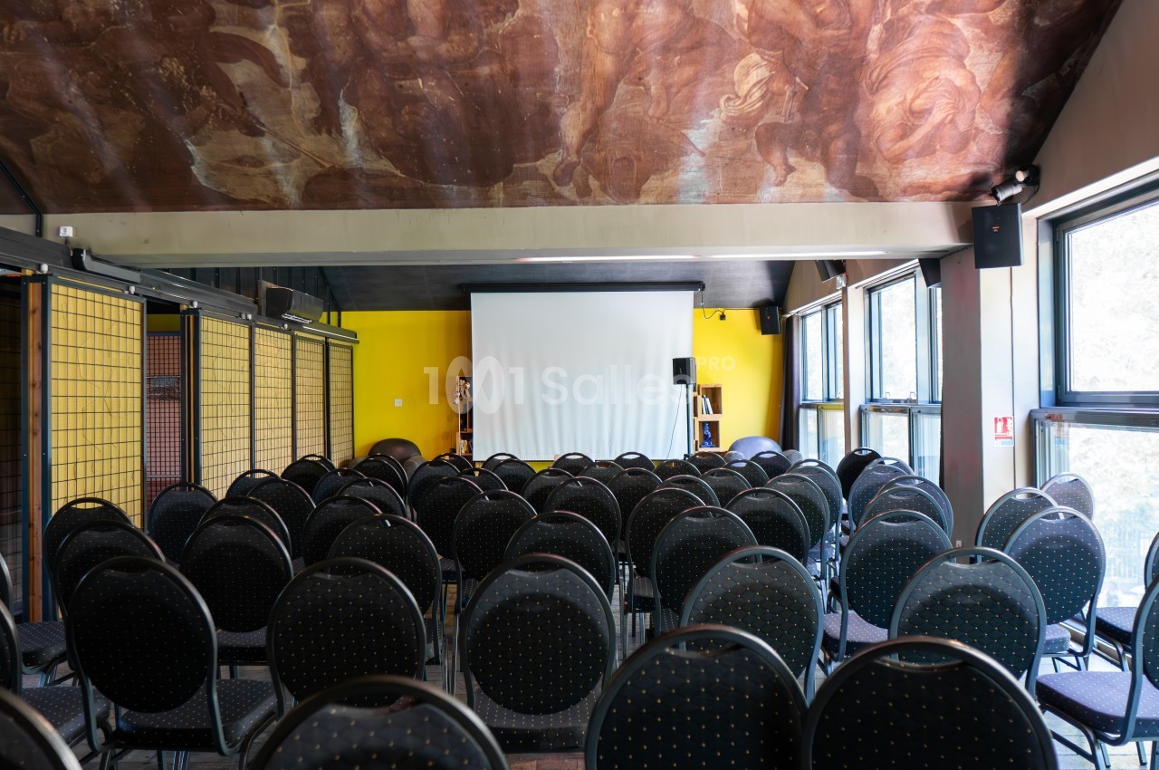 Salle de conférence vide avec des rangées de chaises noires face à un écran de projection blanc.