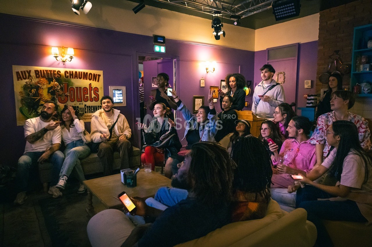 Un groupe de personnes assises et debout dans un salon, regardant un écran hors champ dans une ambiance conviviale.