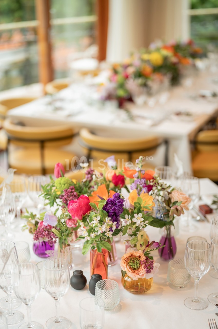 Tables élégamment dressées avec des bouquets de fleurs colorées, des verres et de la vaisselle dans une salle lumineuse.