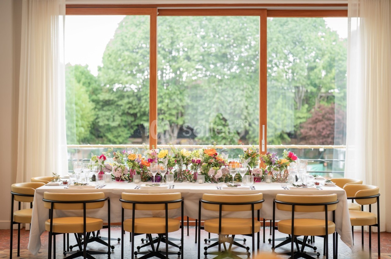 Table dressée avec des fleurs colorées, vaisselle élégante et chaises jaunes, devant une grande baie vitrée donnant sur un…