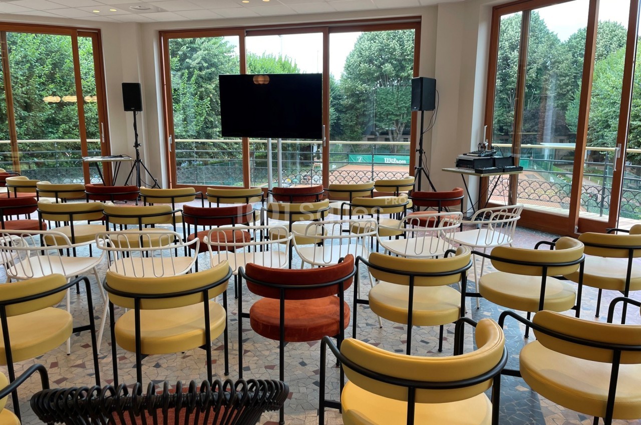 Salle de conférence lumineuse avec des chaises colorées, un écran noir et une vue sur un jardin à travers de grandes…