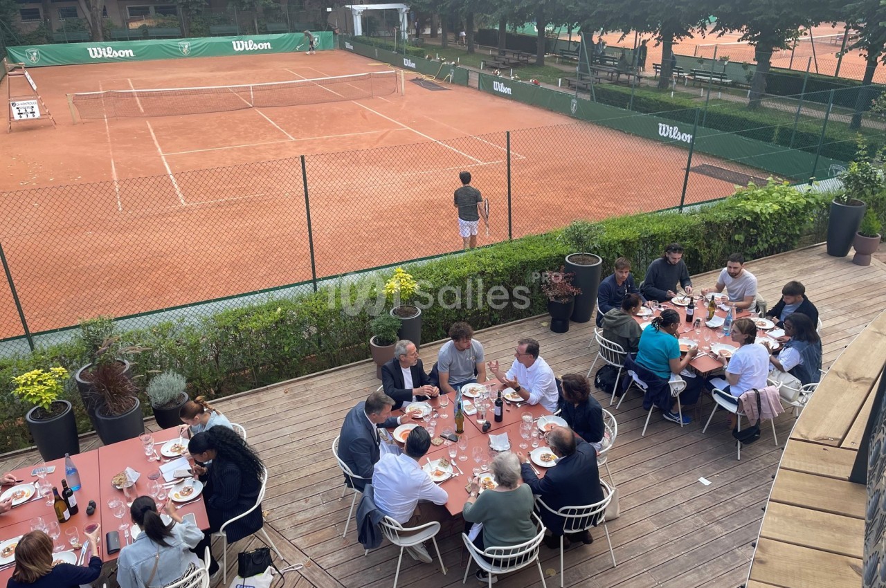 Des personnes dînent sur une terrasse en bois surplombant des courts de tennis en terre battue.
