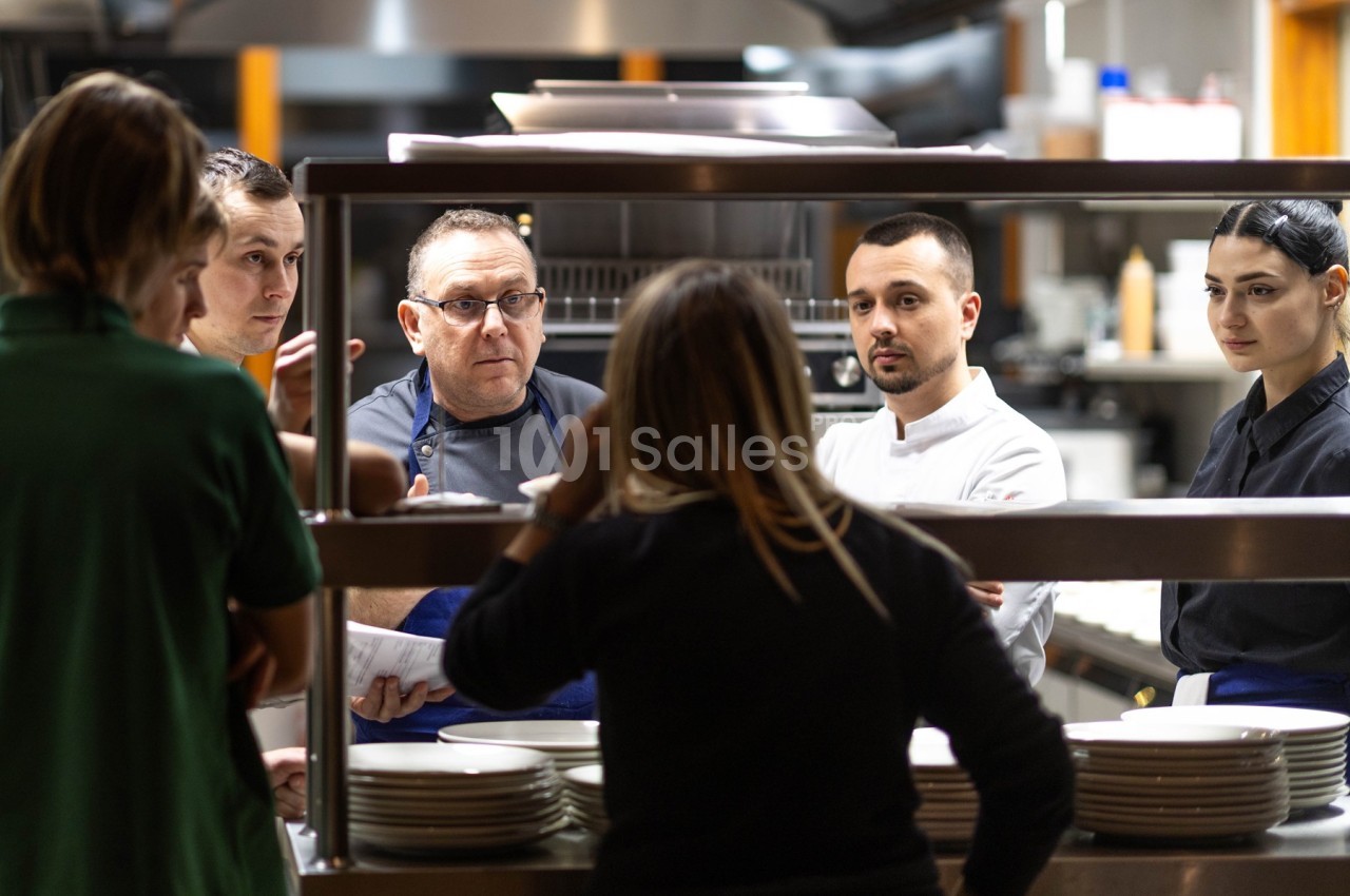 Un groupe de cuisiniers et une femme discutent dans une cuisine professionnelle près d'un passe-plat.