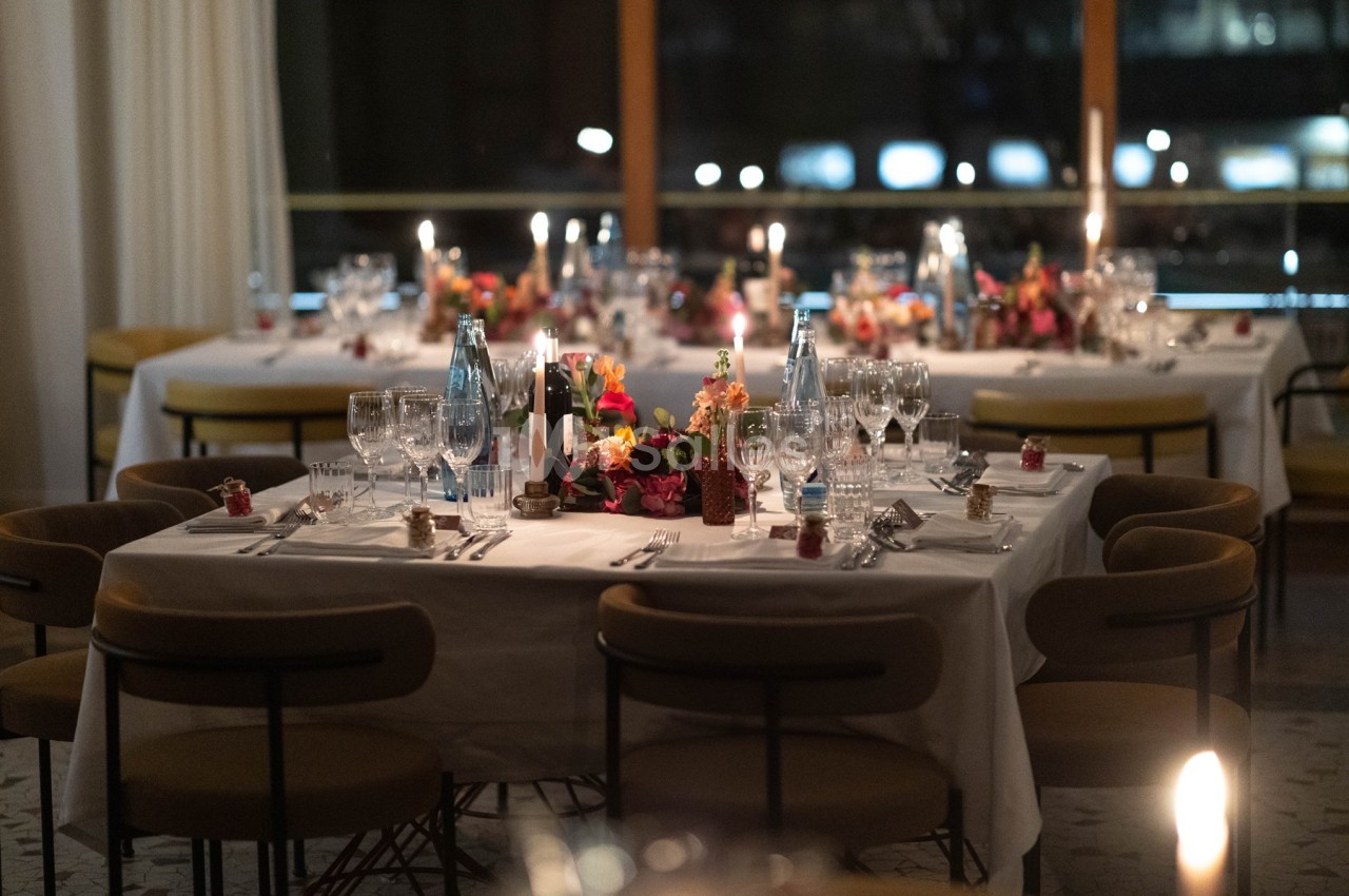Tables élégamment dressées avec nappes blanches, bougies allumées et décorations florales dans une salle éclairée.
