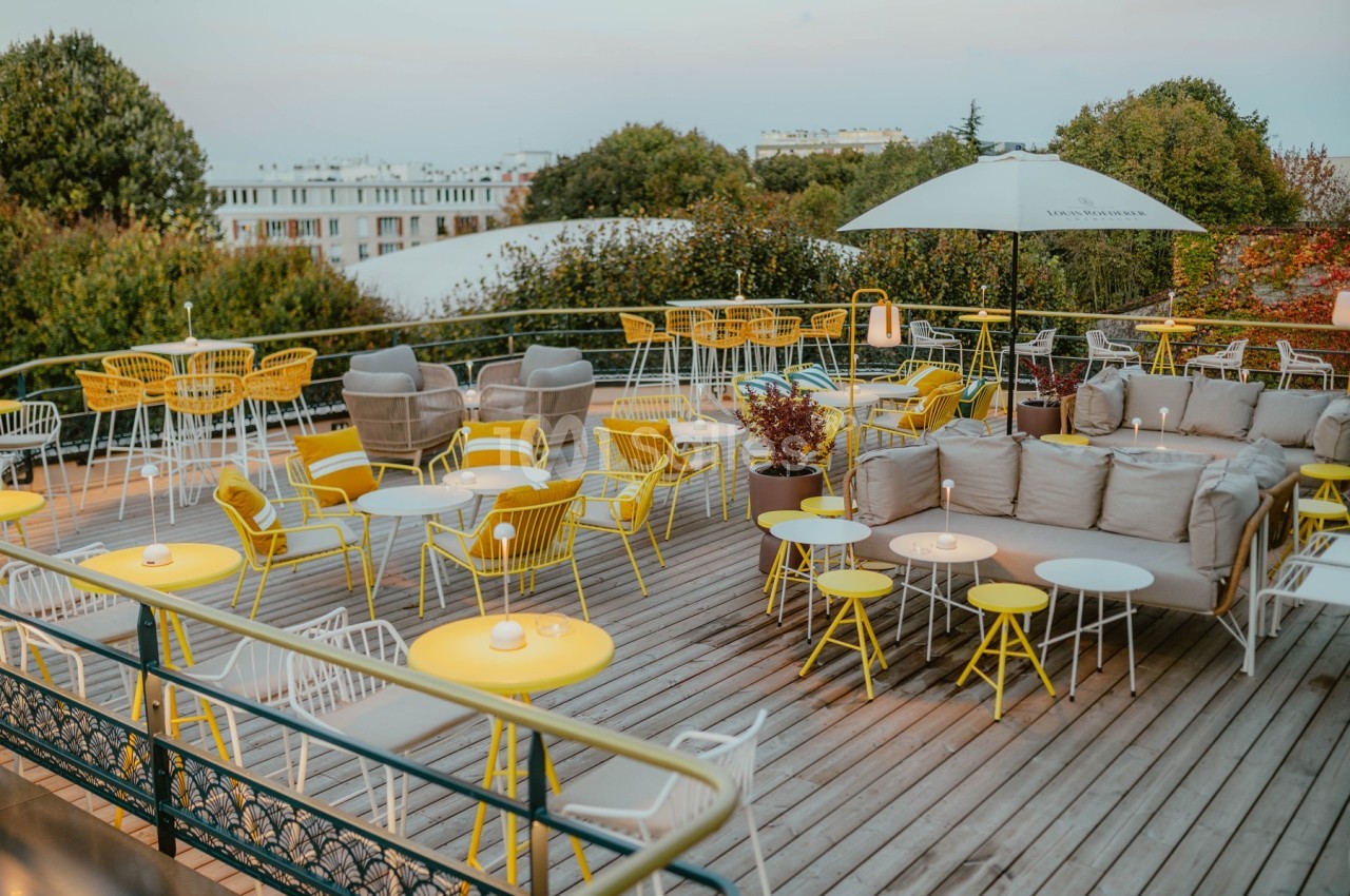 Terrasse en bois aménagée avec des tables et chaises jaunes, des canapés gris et un parasol, entourée de verdure.