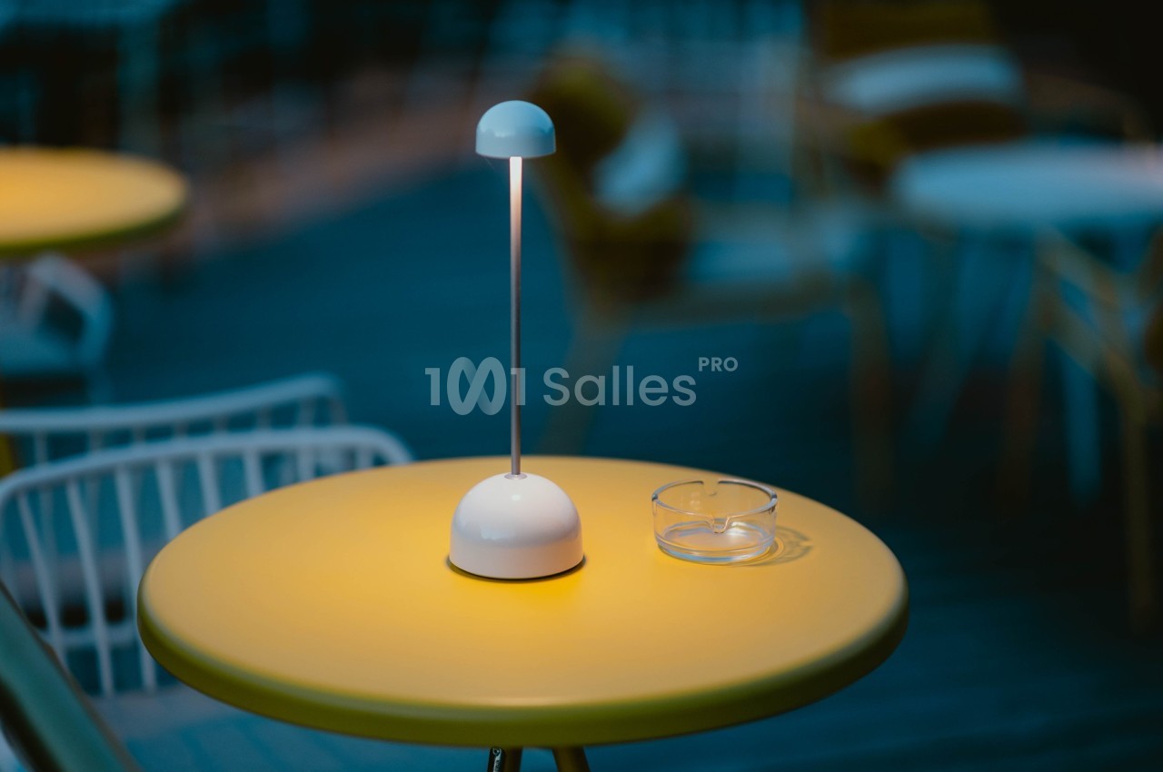 Lampe de table allumée sur une table jaune avec un cendrier en verre, en extérieur, ambiance nocturne floutée.