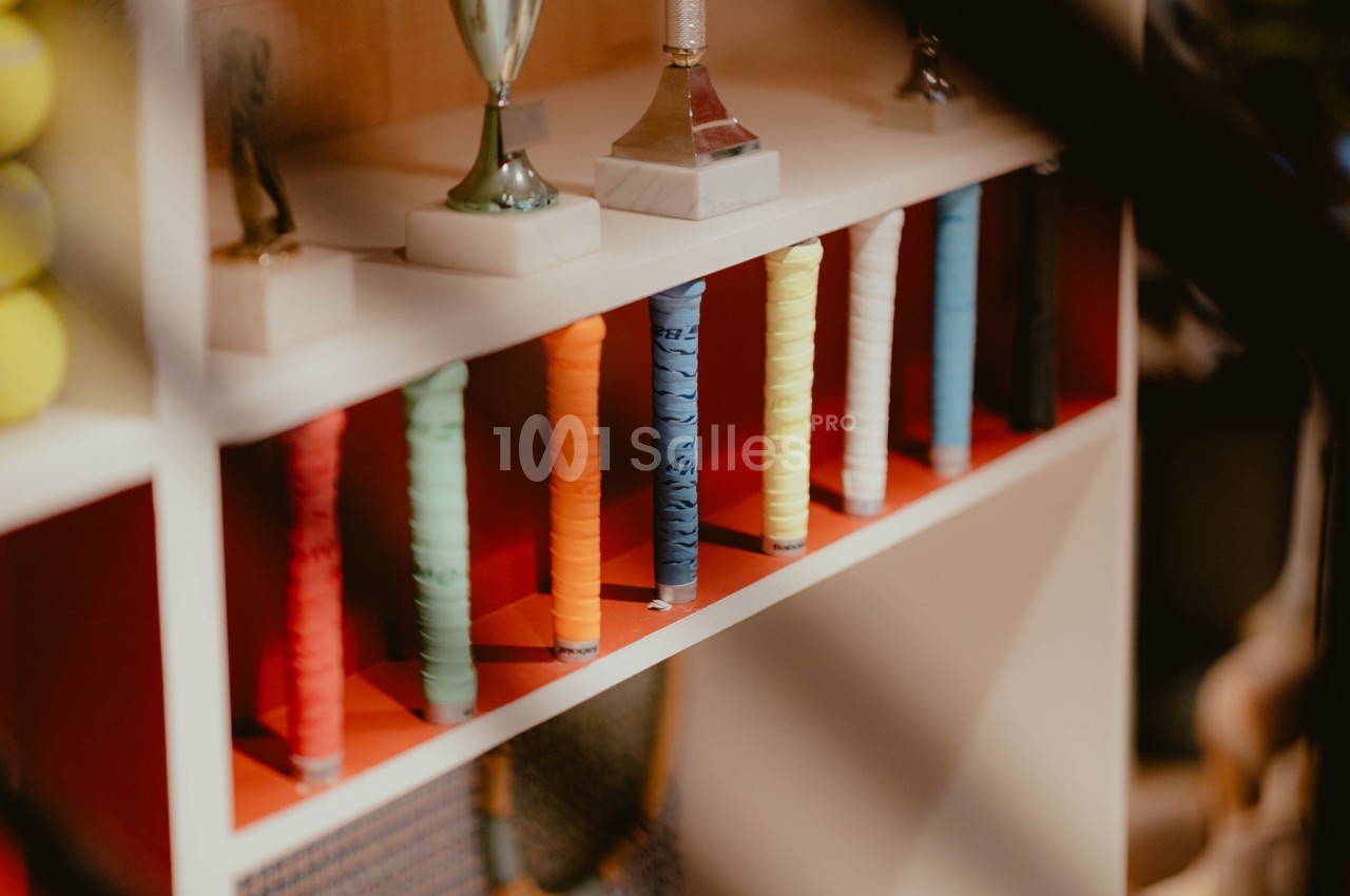 Raquettes de tennis avec poignées colorées alignées sur une étagère, entourées de trophées et d'autres objets.