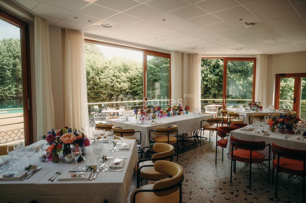 Salle lumineuse avec tables dressées pour un repas, décorées de fleurs colorées, donnant sur une terrasse et un jardin.