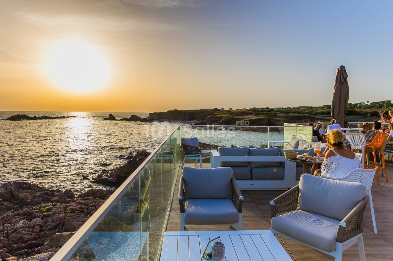 Terrasse en bord de mer au coucher du soleil, avec des tables occupées et des fauteuils vides au premier plan.