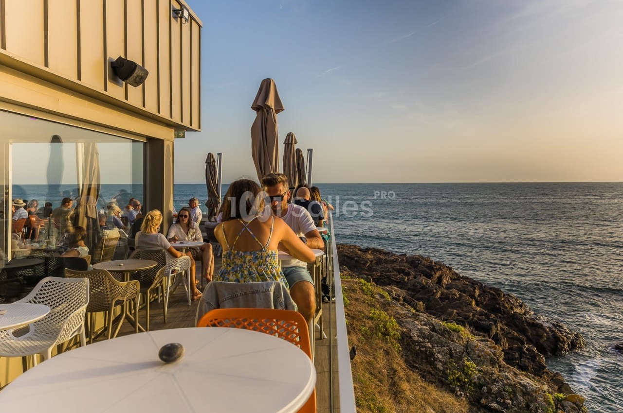 Des personnes assises en terrasse surplombant la mer au coucher du soleil, avec des parasols fermés en arrière-plan.