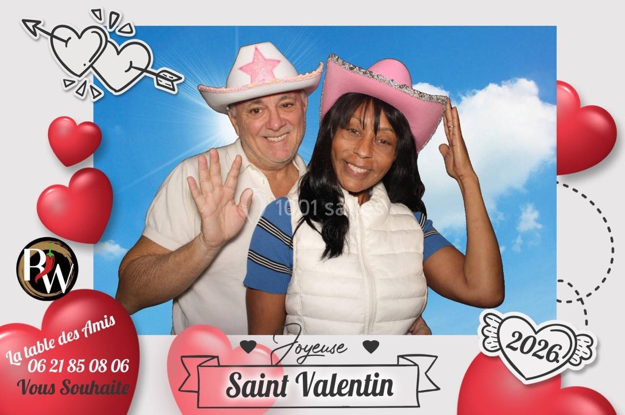 Un couple souriant portant des chapeaux roses pose devant un fond bleu avec des décorations de Saint-Valentin.