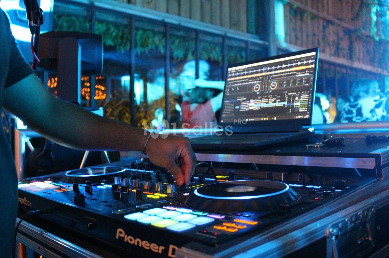 Un DJ manipule une table de mixage éclairée, avec un ordinateur affichant un logiciel de mixage en arrière-plan.