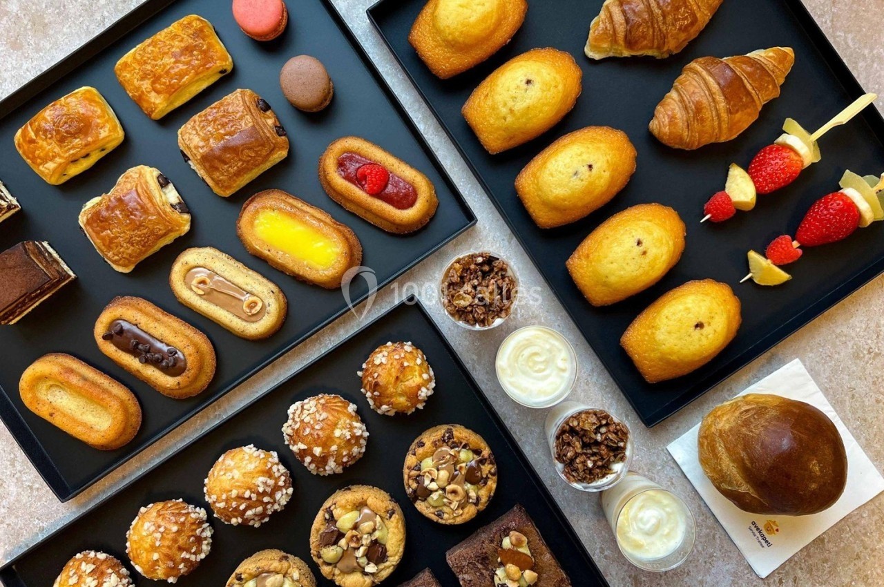 Assortiment de viennoiseries, pâtisseries, muffins, madeleines et desserts présentés sur des plateaux noirs.