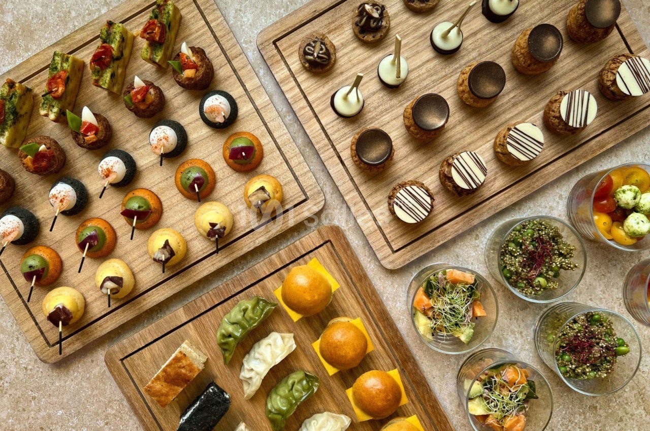 Plateaux en bois garnis de bouchées salées et sucrées, accompagnés de verrines colorées sur une table beige.