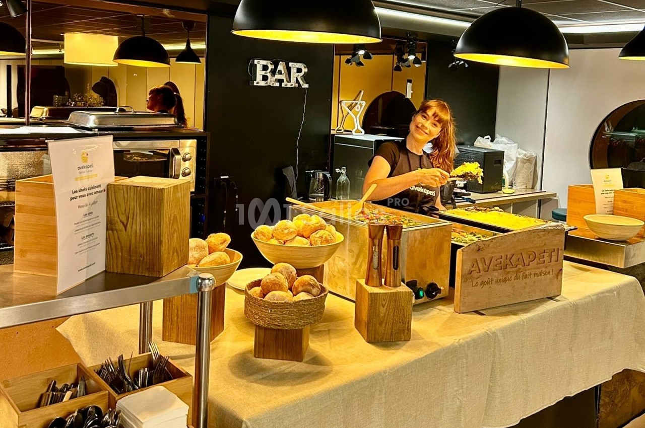 Buffet avec pains, plats variés et une femme souriante servant de la nourriture dans un espace éclairé.