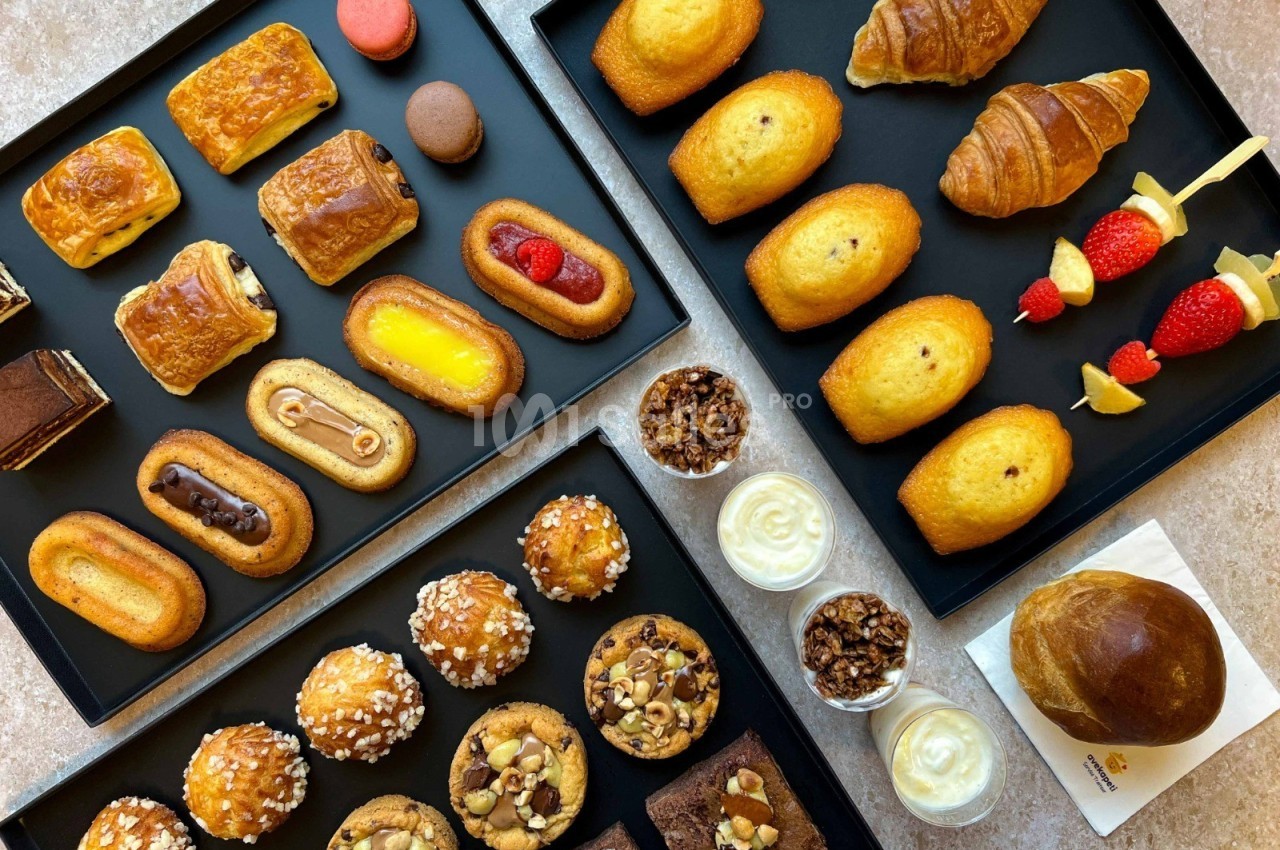 Assortiment de viennoiseries, pâtisseries, madeleines, muffins et desserts disposés sur des plateaux noirs.