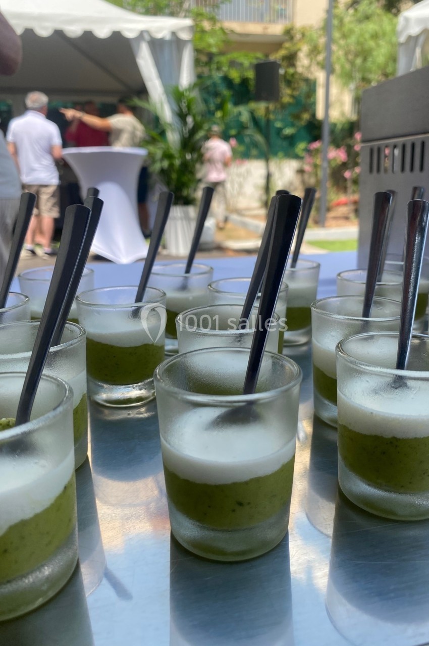 Verrines de soupe froide verte surmontées d'une mousse blanche, disposées sur une table en extérieur.