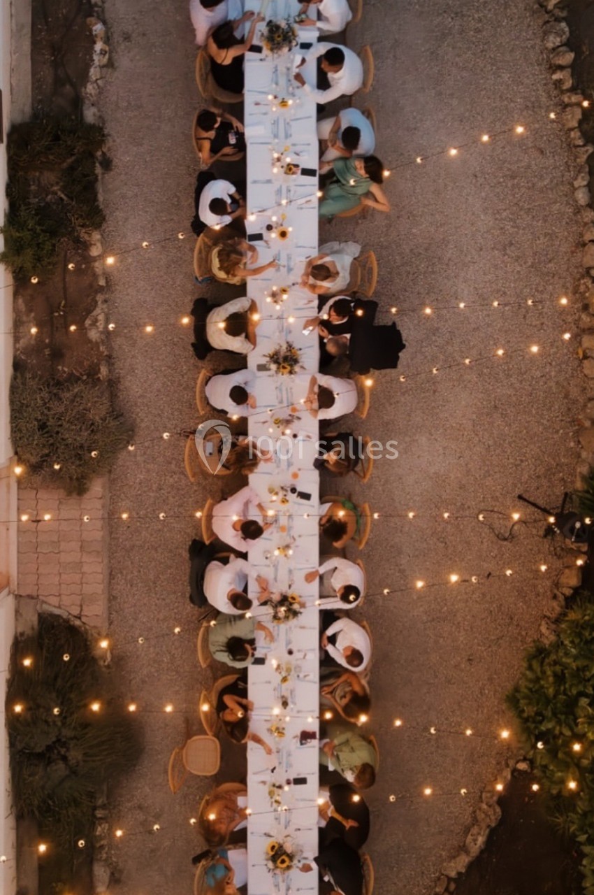 Vue aérienne d'une longue table dressée en extérieur, entourée de convives et éclairée par des guirlandes lumineuses.