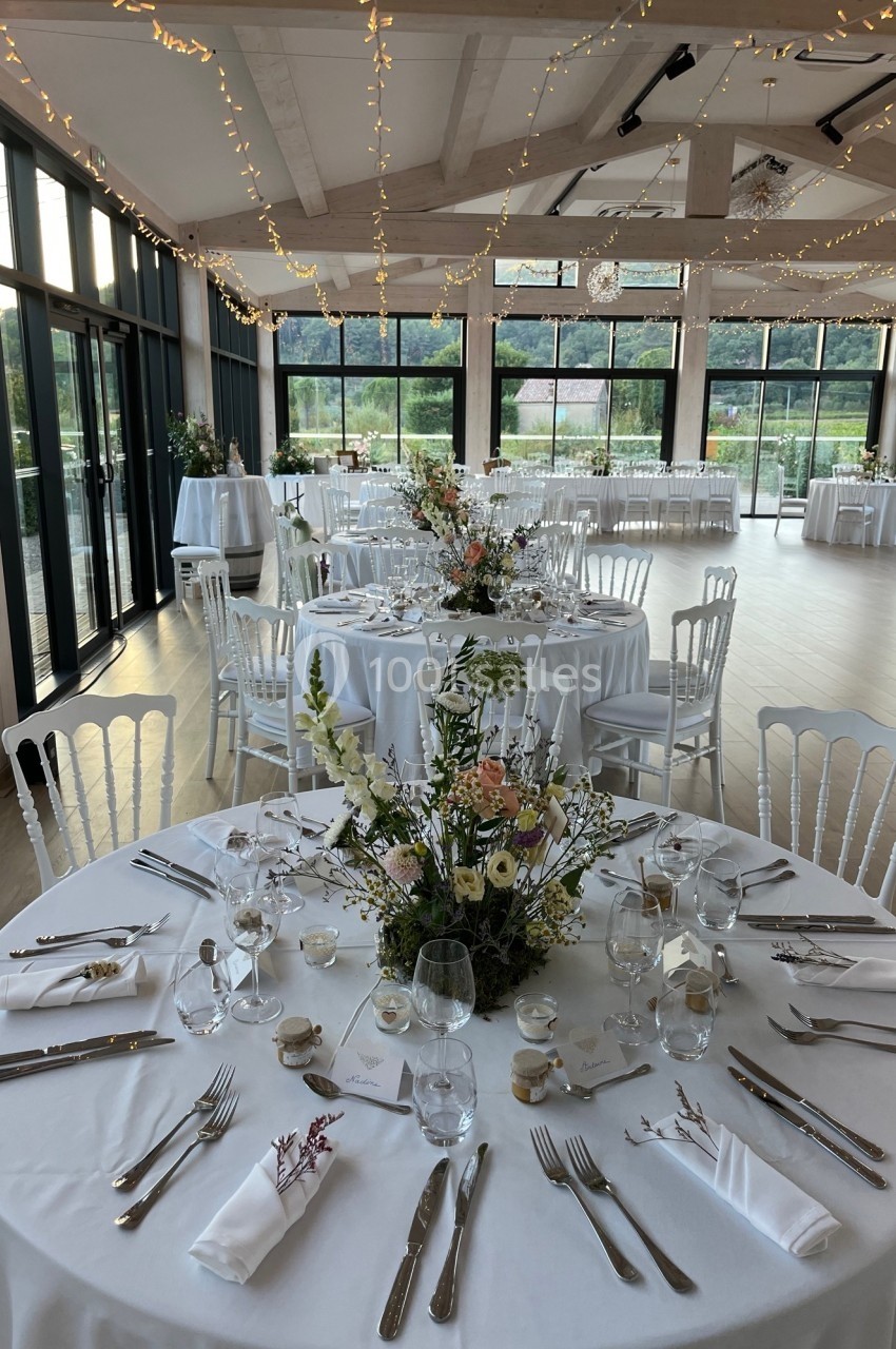 Salle de réception lumineuse avec tables rondes décorées de fleurs et vaisselle élégante, éclairée par des guirlandes.