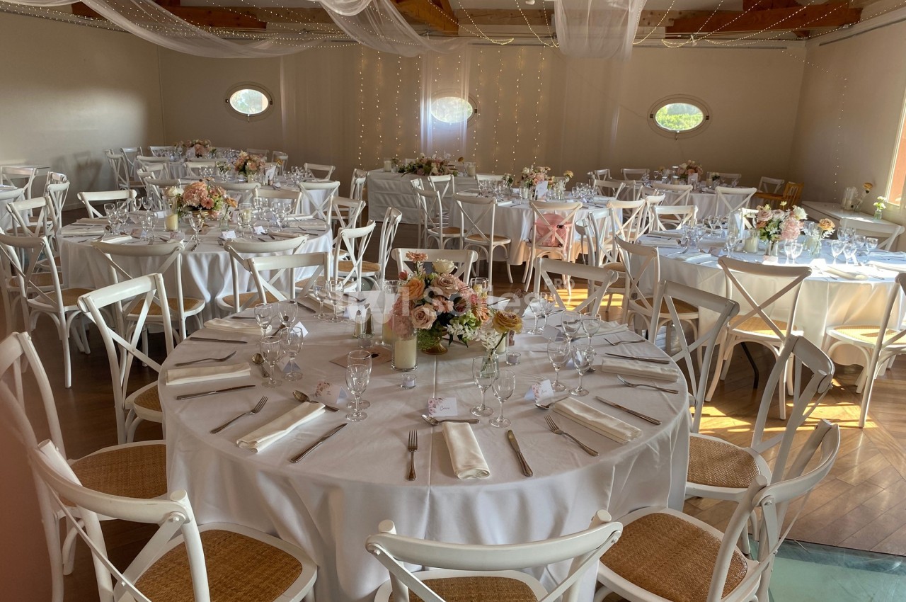 Salle de réception décorée pour un mariage, avec tables rondes dressées, nappes blanches et arrangements floraux.