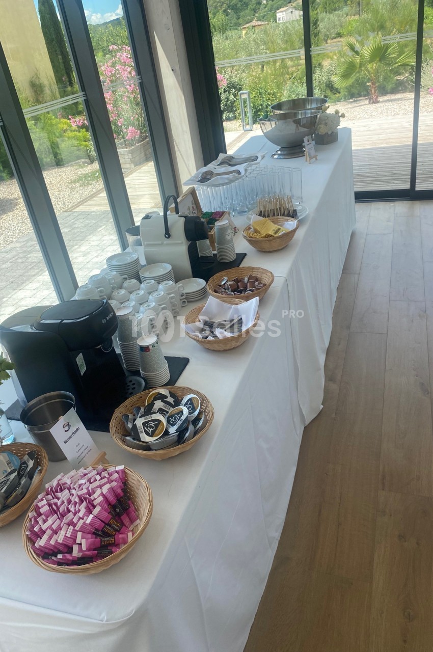 Table dressée avec boissons chaudes, viennoiseries et condiments, située près de grandes baies vitrées donnant sur un jardin.