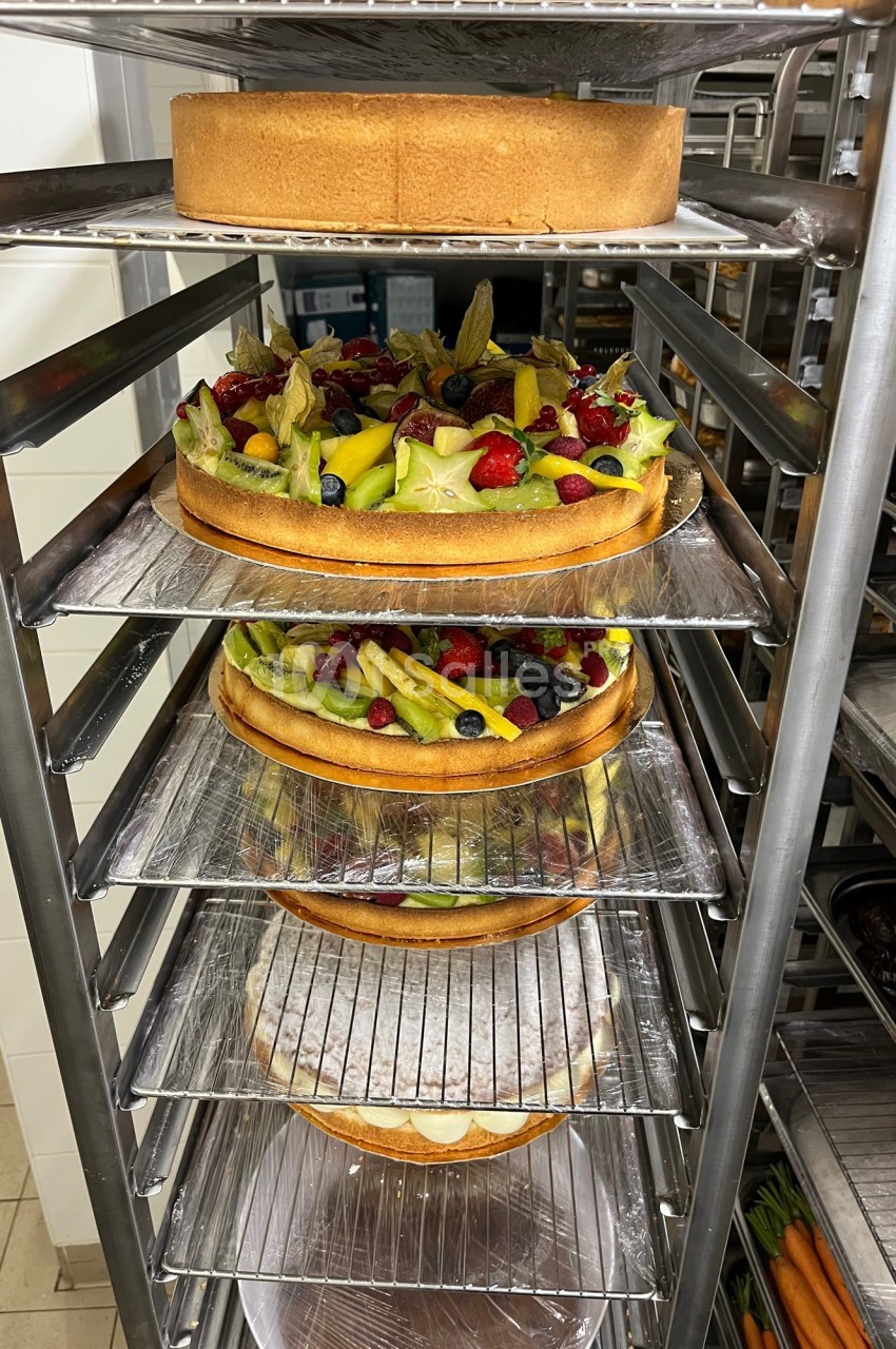 Gâteaux garnis de fruits frais et tartes sur des étagères métalliques dans une cuisine professionnelle.
