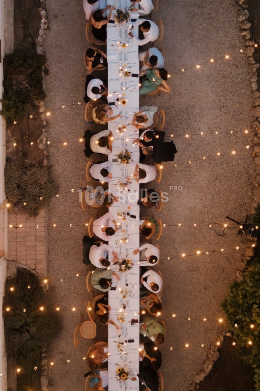 Vue aérienne d'une longue table dressée en extérieur, entourée de convives et éclairée par des guirlandes lumineuses.