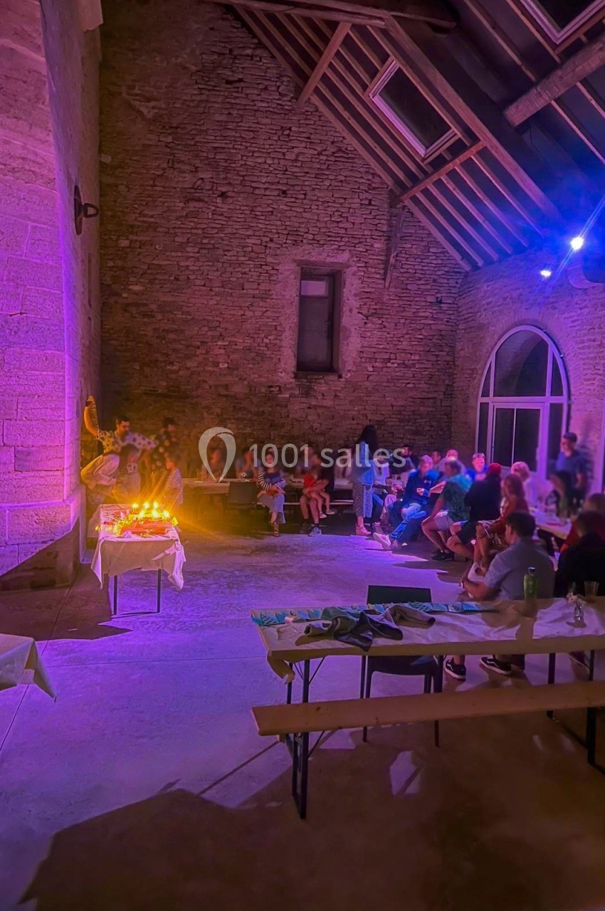 Groupe de personnes réunies dans une salle en pierre éclairée par des lumières colorées, avec des tables et des bougies.