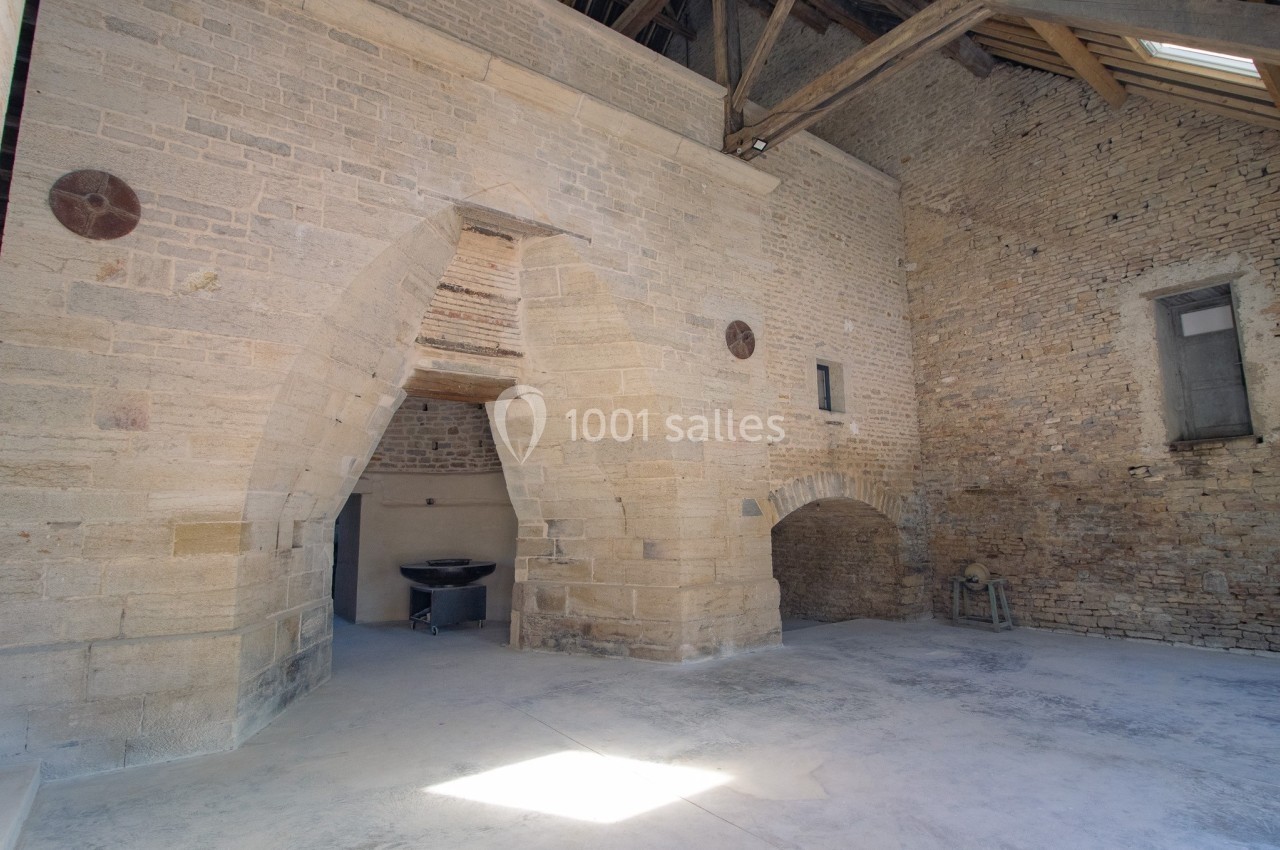 Intérieur d'un bâtiment en pierre avec des arches, un sol en béton et une charpente en bois visible au plafond.