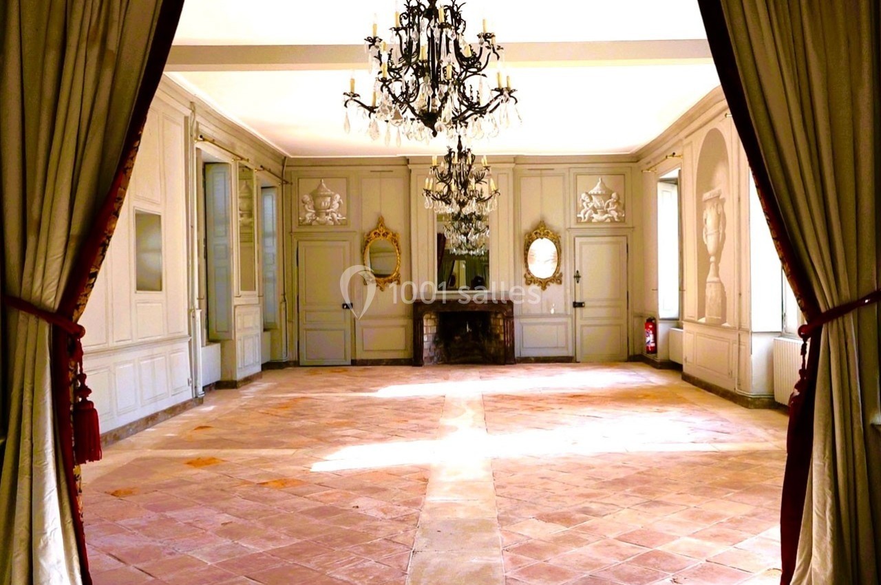 Salle lumineuse avec sol en carreaux de terre cuite, murs clairs ornés de moulures et lustre en cristal suspendu.