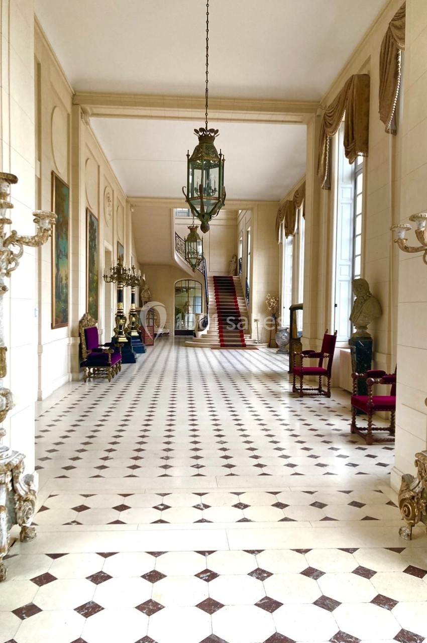 Hall spacieux avec sol en damier, chandeliers, fauteuils anciens, escalier central et grandes fenêtres.