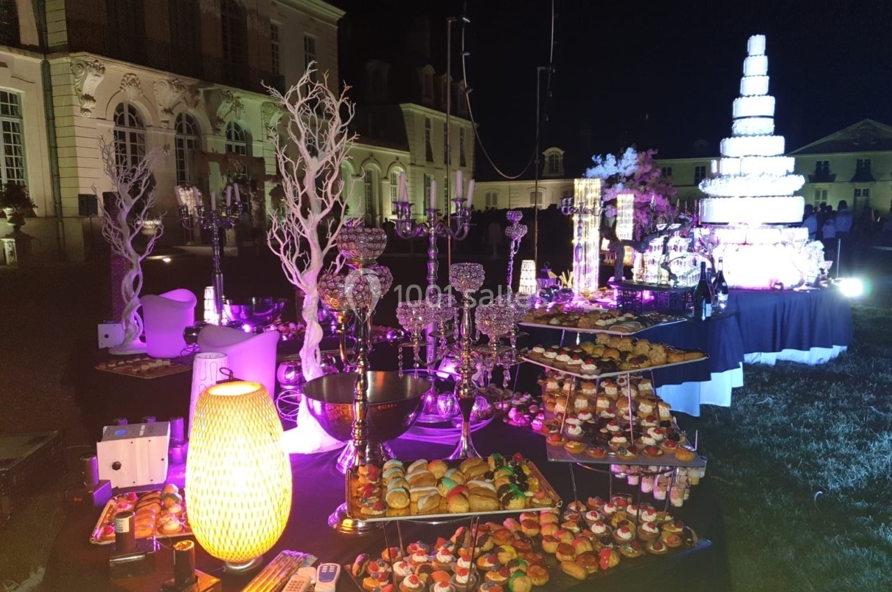 Buffet éclairé en extérieur de nuit, avec desserts variés, chandeliers et grande pièce montée illuminée en arrière-plan.